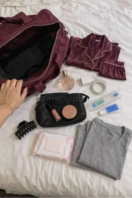 An open maroon overnight bag on a white hotel bed with a satin night set, perfume, charger, toothbrush, makeup pouch, hair clip, face wipes and a grey T-shirt